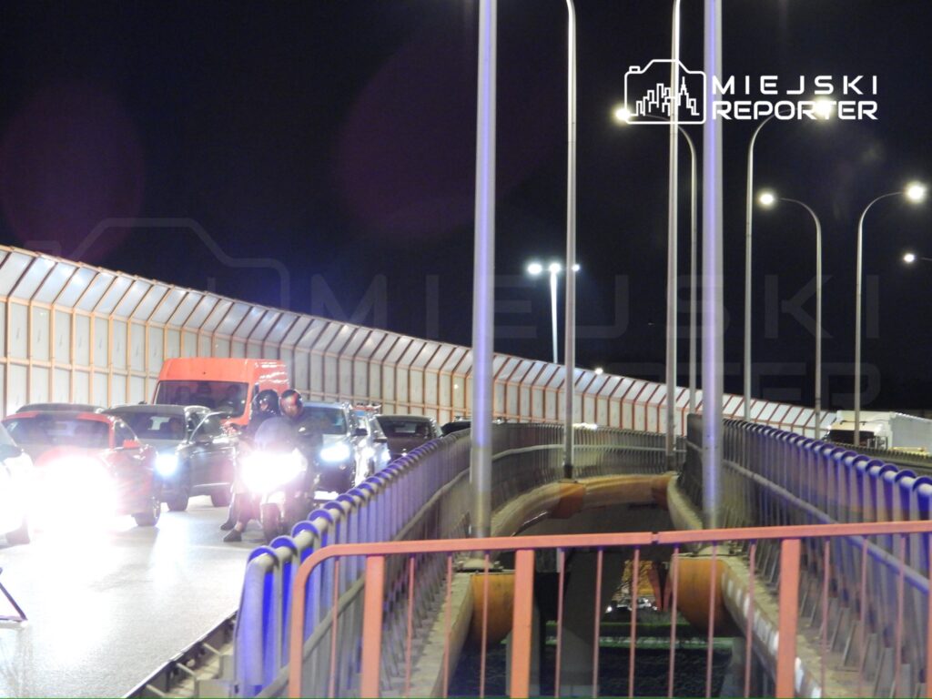 Ruch samochodowy i motocyklowy na oświetlonym moście w nocy, z widoczną osłoną z tworzywa sztucznego.