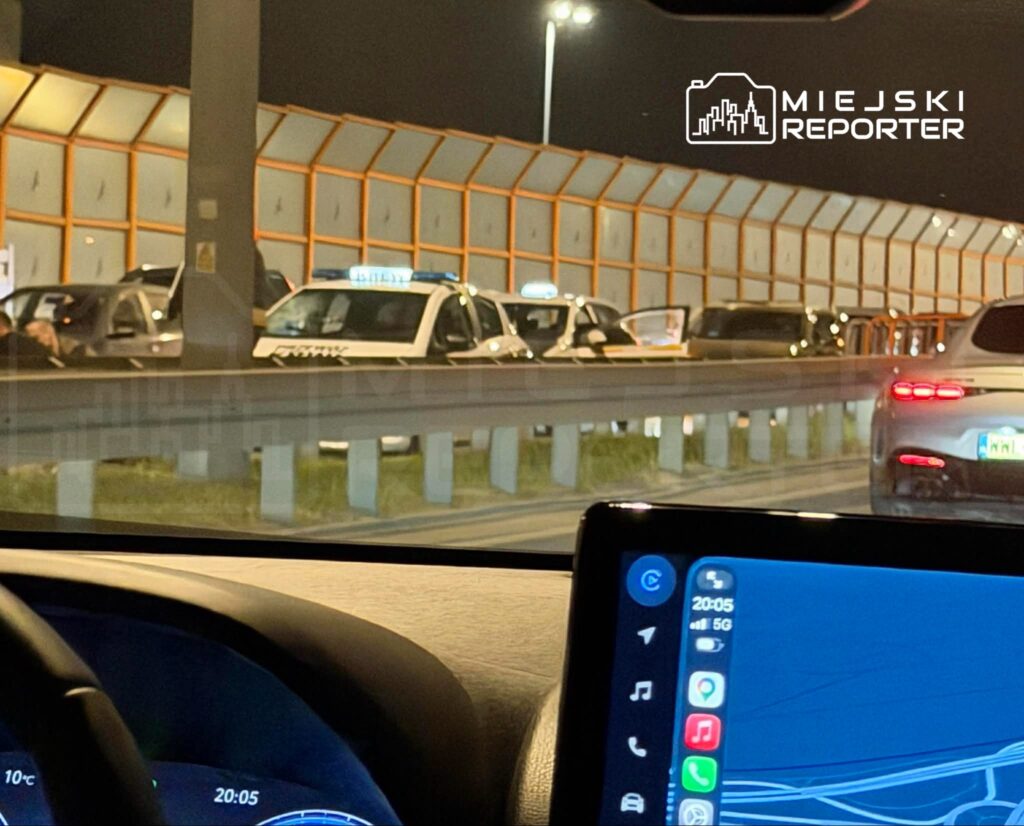 Ruch samochodowy zatrzymany w nocy, w tle widoczne radiowozy oraz inne pojazdy na drodze.