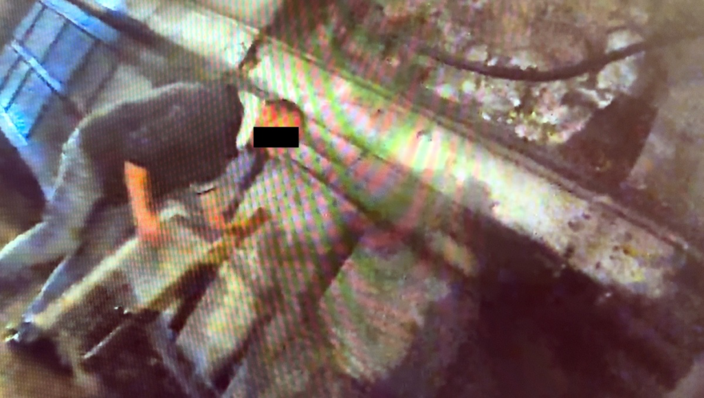 Mężczyzna w ciemnym ubraniu pochyla się nad ziemią w słabo oświetlonym pomieszczeniu.