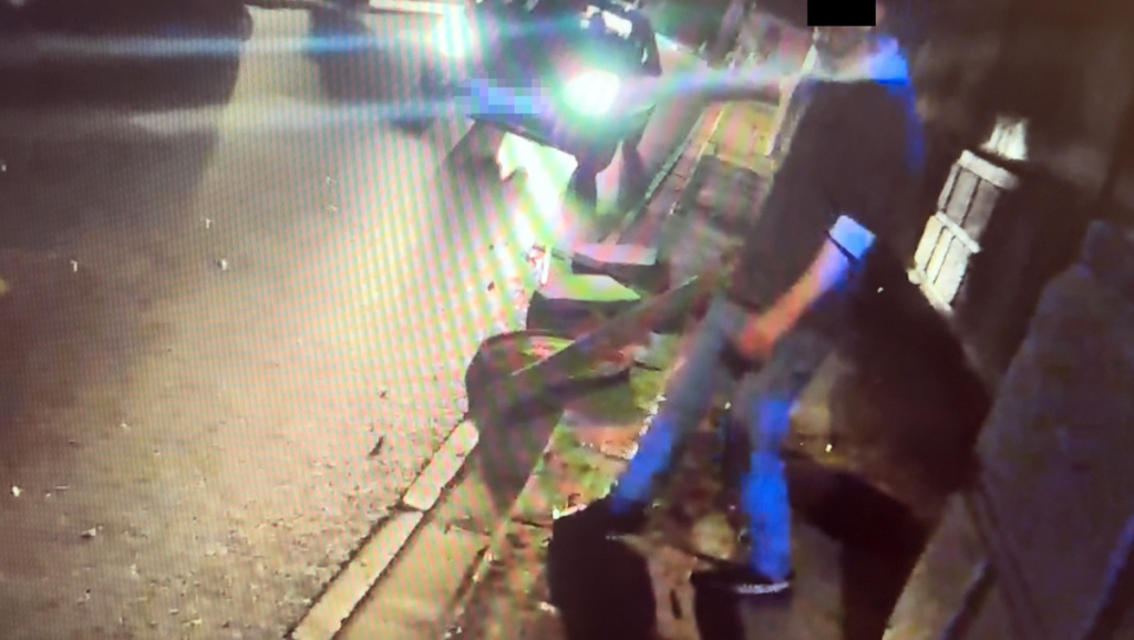 Mężczyzna w ciemnym ubraniu stoi na chodniku, obok przejeżdżającego samochodu z włączonymi światłami.