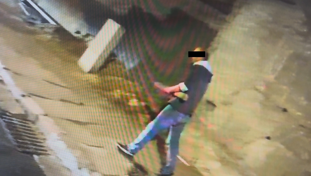 Mężczyzna w ciemnej odzieży idący po mokrej nawierzchni, w tle widoczna ściana budynku.