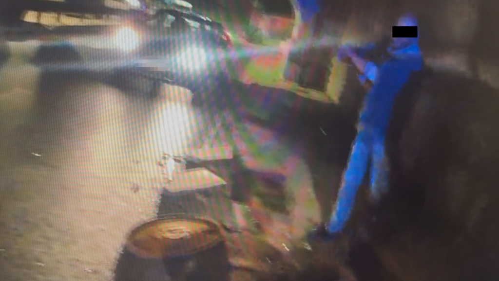 Mężczyzna w niebieskiej koszuli stoi obok pojazdu, trzymając coś w rękach, na ciemnej ulicy oświetlonej reflektorami.