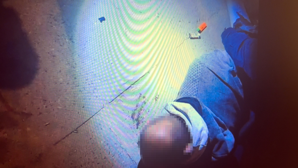 Mężczyzna leżący na ziemi w szarej bluzie, obok widoczny przedmiot w pomarańczowej obudowie.