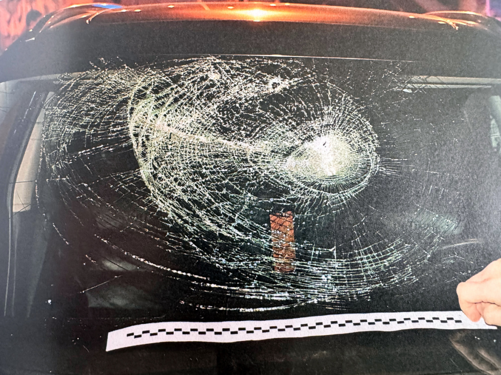 Pęknięta tylna szyba samochodu z wyraźnymi śladami uderzenia, obok ręka trzymająca taśmę pomiarową.