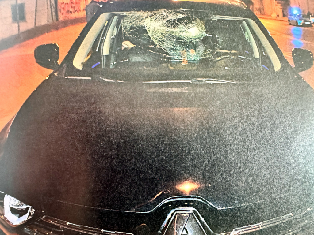 Czarny samochód z pękniętą szybą przednią, stojący na oświetlonej ulicy. W tle widać niebieskie światła radiowozu.