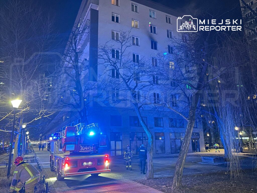 W nocy strażacy przy wozie bojowym interweniują przed budynkiem mieszkalnym, w otoczeniu drzew i lamp oświetleniowych.