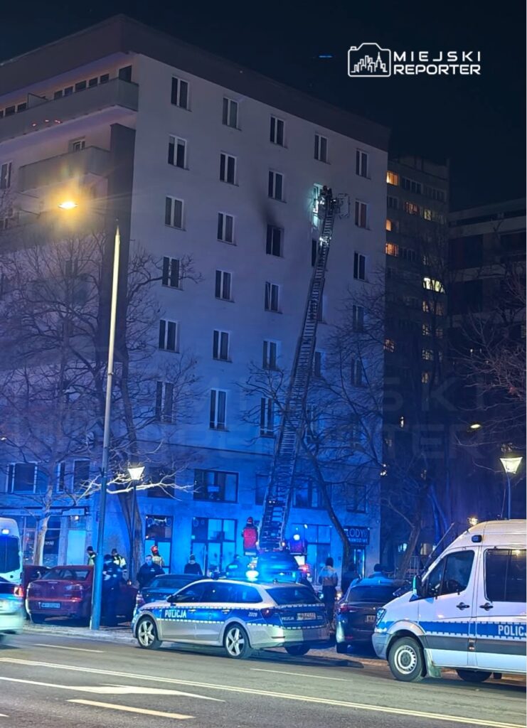 Strażacy na drabinie ratują osobę z okna na wyższym piętrze budynku w nocy, obok pojazdy policyjne i karetki.