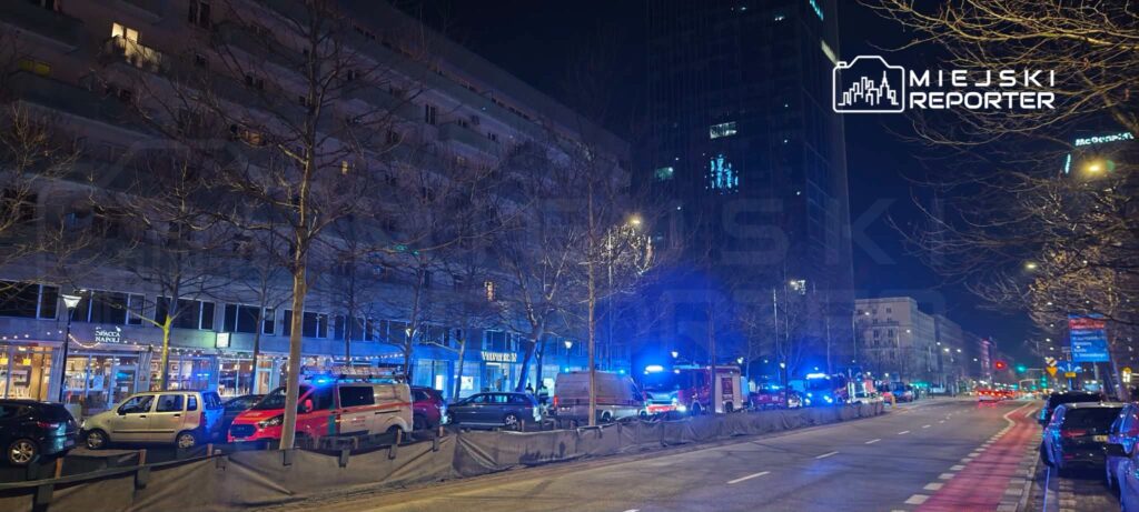 Nocna scena z policyjnymi i strażackimi pojazdami zaparkowanymi wzdłuż ulicy, oświetlona niebieskimi światłami.