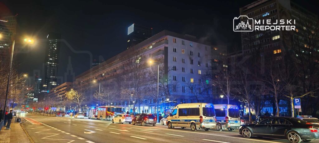 Ulica nocą z widocznymi pojazdami policyjnymi i strażackimi, otoczona nowoczesnymi budynkami i drzewami.