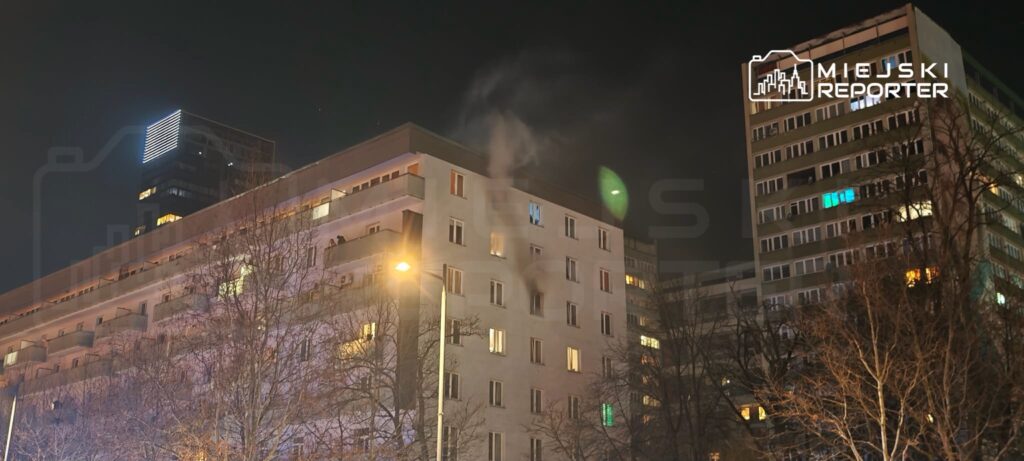 Dwie osoby stojące na balkonie, w tle nowoczesne budynki oświetlone nocą, z dymem wydobywającym się z komina.