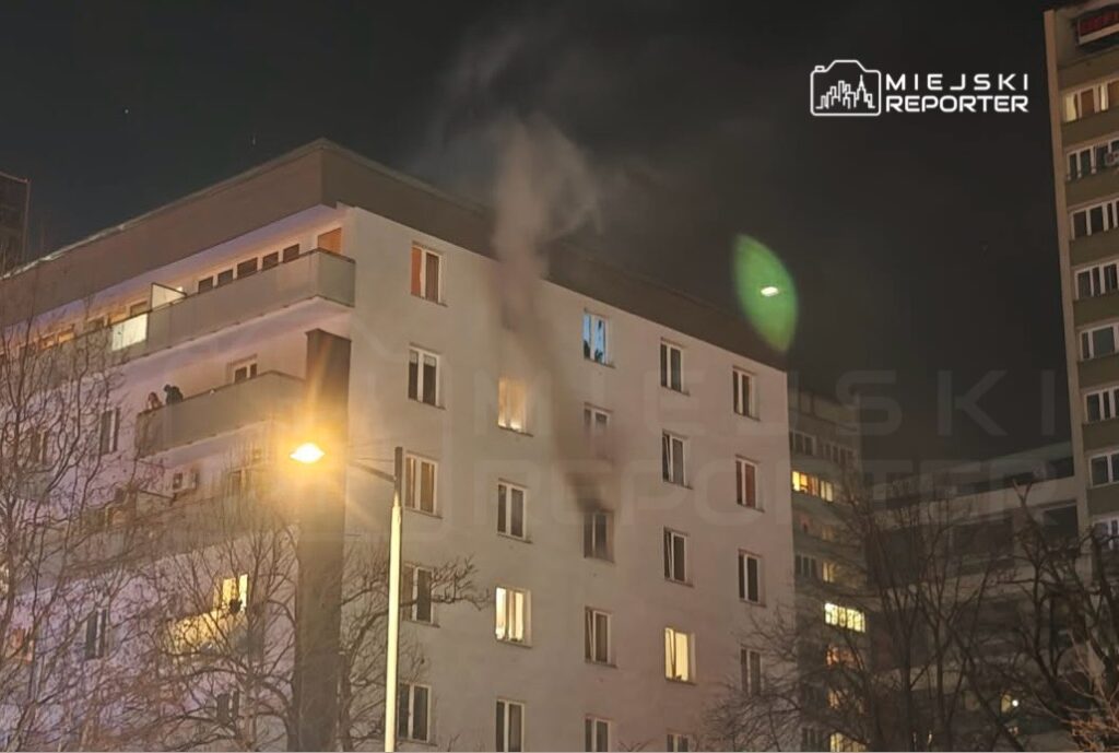 Dym wydobywający się z okna mieszkania w bloku mieszkalnym w nocy, w tle widoczne inne budynki.