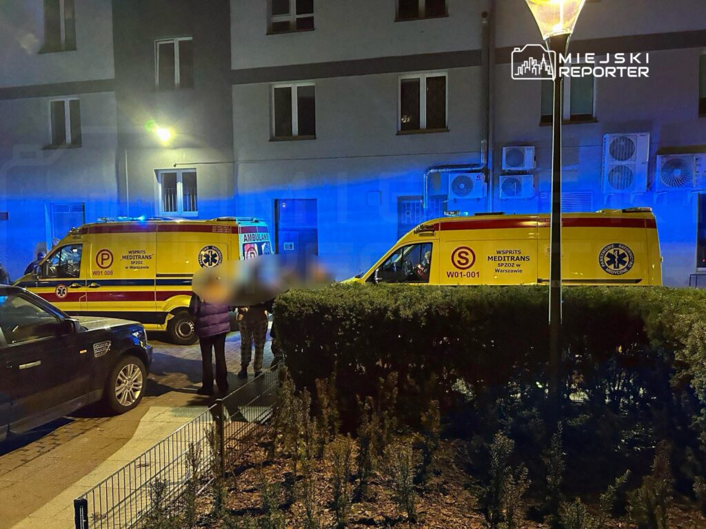 Karetka pogotowia z włączonymi sygnałami świetlnymi stoi na ulicy, obok grupy ludzi w rozmowie.