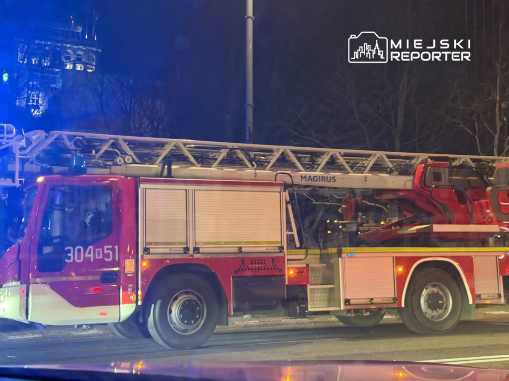 Czerwony wóz strażacki z drabiną, oświetlony niebieskimi sygnalizatorami, stoi na ulicy nocą.