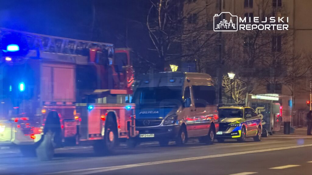 W nocy na ulicy widać pojazdy służb ratunkowych, w tym wóz strażacki i radiowóz policyjny z włączonymi sygnałami świetlnymi.