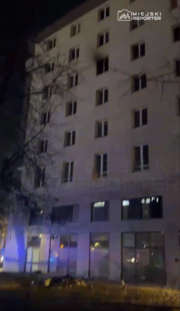 Nowoczesny budynek mieszkalny z otwartymi oknami, oświetlony niebieskimi światłami policyjnymi w nocy.