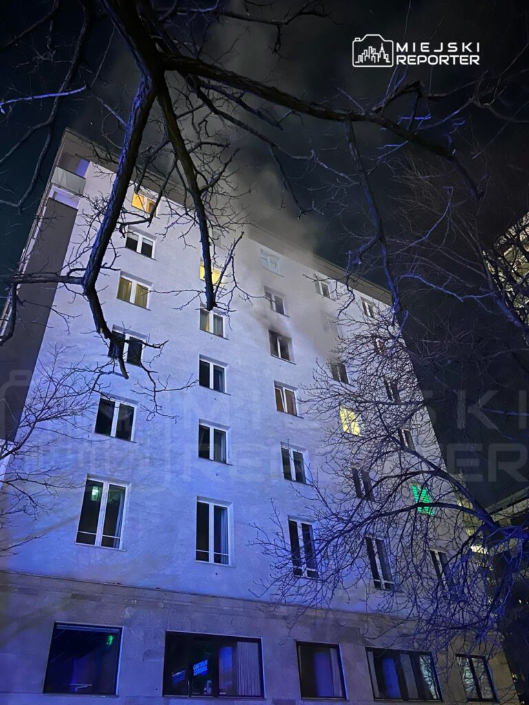 Widok na nowoczesny budynek mieszkalny z dymem wydobywającym się z okien, otoczony nagimi gałęziami drzew w nocy.