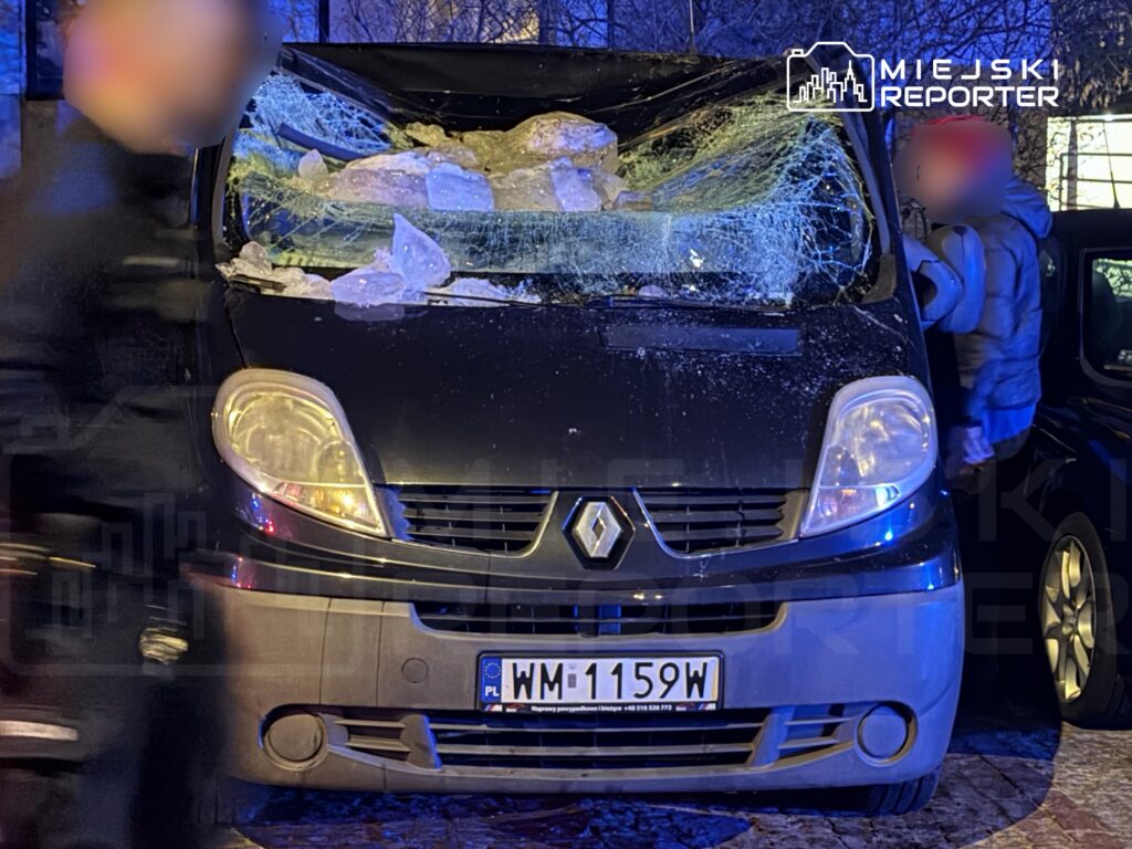 Zniszczony van z rozbitą szybą i odłamkami lodu na dachu, zaparkowany na ulicy w nocy.