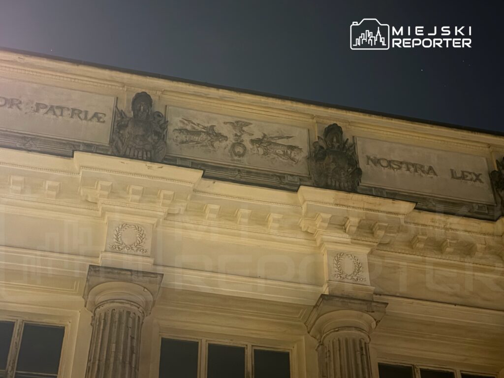 Fasada budynku z rzeźbami i inskrypcjami "PATRIAE NOSTRA LEX" oświetlona nocnym światłem.