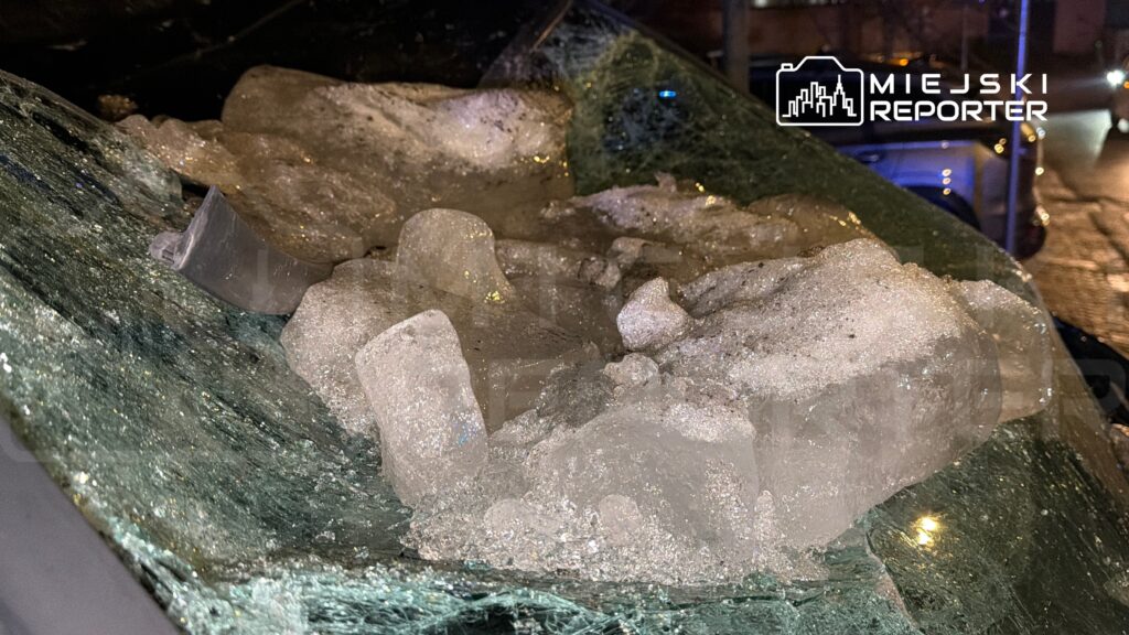 Szkło samochodowe pokryte dużymi bryłami lodu, z widocznymi rysami i pęknięciami, w nocnym otoczeniu miejskim.