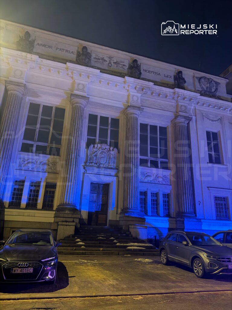 Neoklasycystyczny budynek z kolumnami oświetlony niebieskim światłem, z zaparkowanymi samochodami na chodniku.