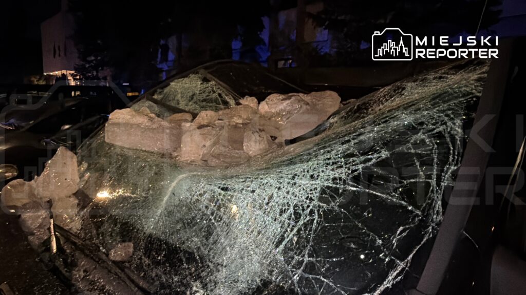 Samochód z rozbitą szybą, pokryty dużymi bryłami kamieni, zaparkowany w ciemnej ulicy.
