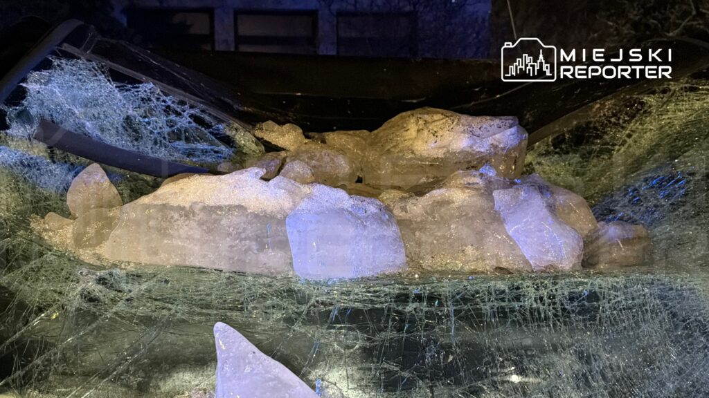 Skały lodu leżą na uszkodzonym samochodzie, otoczone pękniętym szkłem i reflektorami w tle.