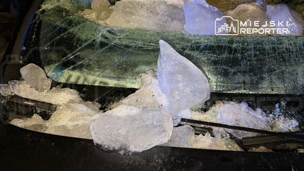 Szyba samochodu pokryta dużymi bryłami lodu, z widocznymi pęknięciami. W tle ciemne otoczenie.