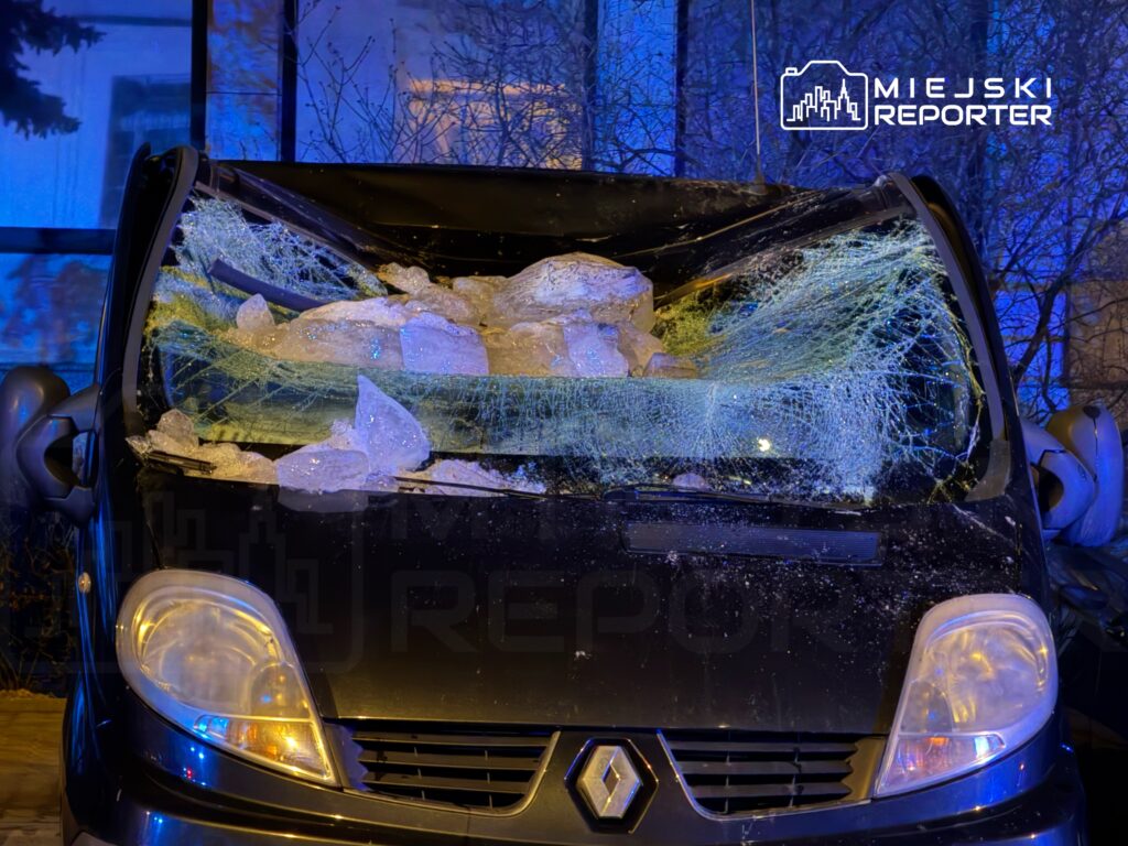 Czarna furgonetka z rozbitą szybą przednią, na której leżą duże kawałki lodu, w nocnym otoczeniu z niebieskim oświetleniem.