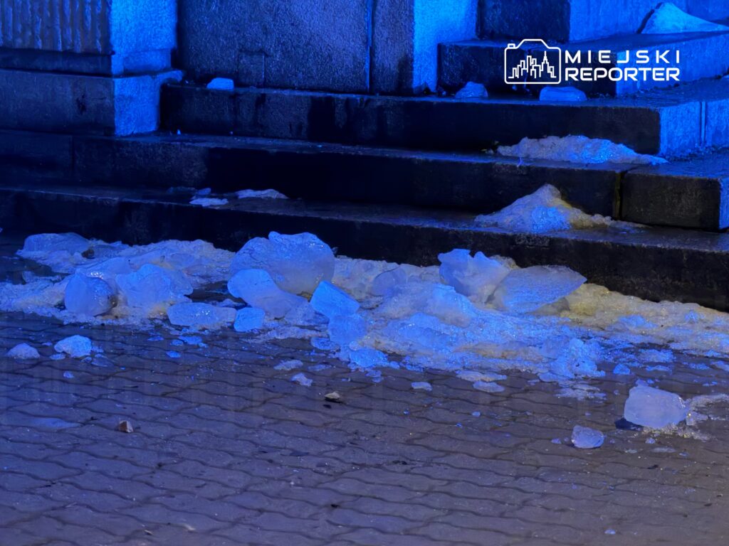 Lodowe bryły leżą na mokrej kostce brukowej pod niebieskim oświetleniem przy schodach.