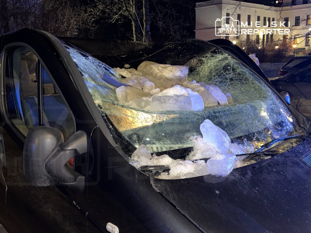 Uszkodzony samochód z rozbitą szybą, na dachu leżą bryły lodu w oświetlonym nocą otoczeniu.