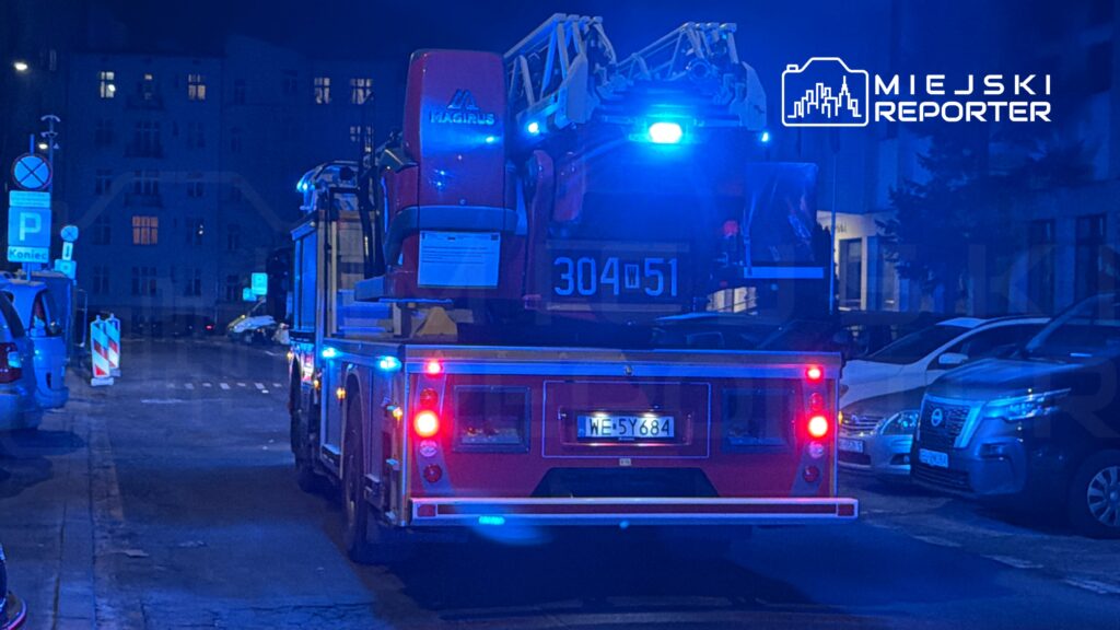 Czerwony wóz strażacki z włączonymi sygnałami świetlnymi stoi na ulicy w nocy, obok zaparkowanych samochodów.