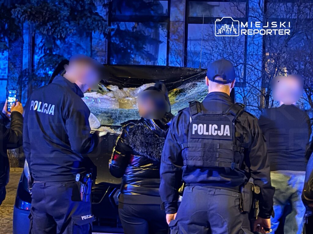 Grupa policjantów w mundurach prowadzi czynności przy uszkodzonym pojeździe, otoczona przez obserwujących.