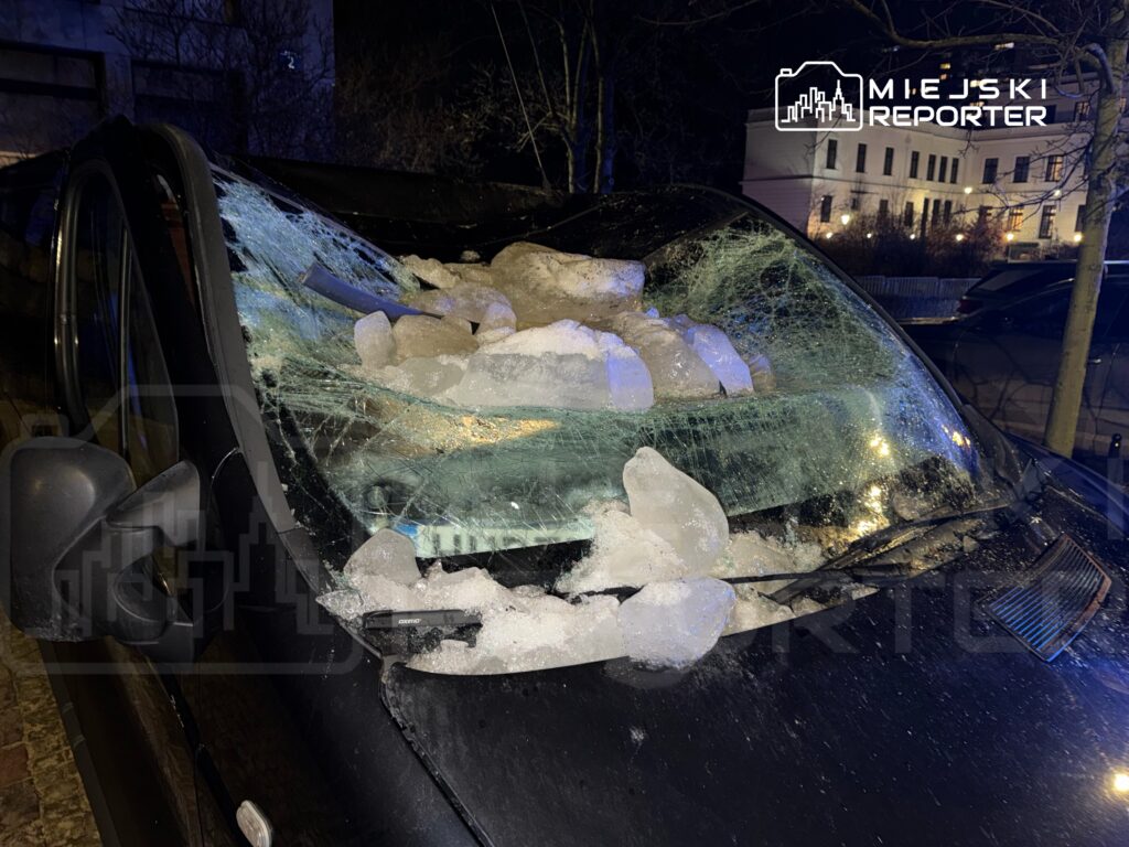Czarna furgonetka z rozbitą szybą, pokrytą lodowymi bryłami, zaparkowana w ciemnej ulicy nocą.