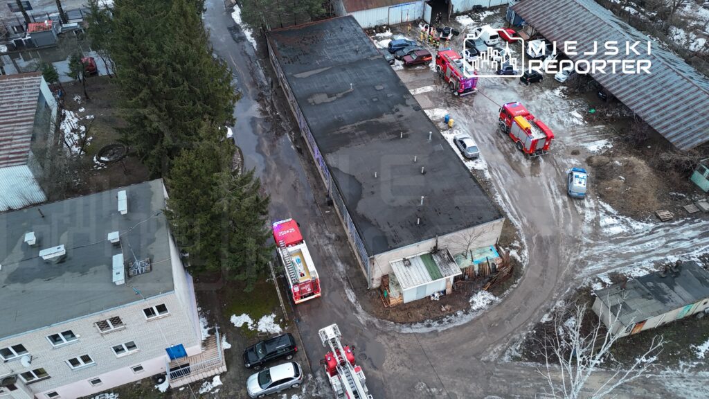 Strażacy w sprzęcie gaśniczym pracują przy budynku przemysłowym na terenie z błotnistą drogą i pozostałościami śniegu.