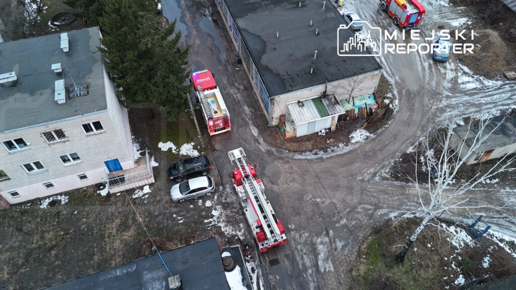 Czerwony wóz strażacki z drabiną stoi na wąskiej ulicy obok budynku, otoczony przez śnieg i błoto.