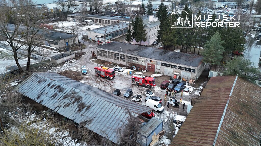 Grupa strażaków oraz ratowników medycznych przy samochodach na terenie przemysłowym z widocznymi budynkami i śniegiem.