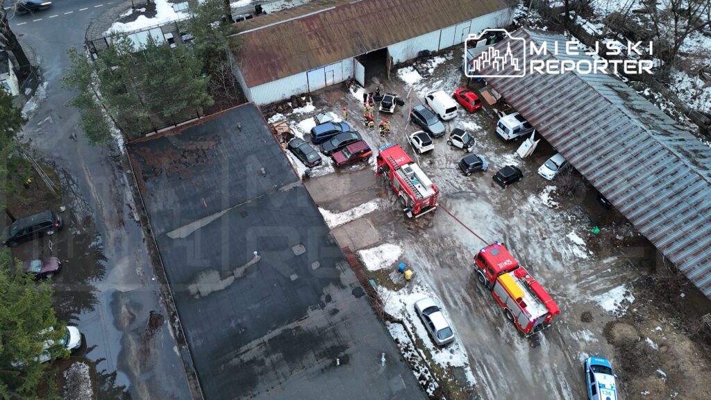 Grupa strażaków pracuje przy pożarze w zakładzie przemysłowym, obok zaparkowane samochody i zniszczony teren.