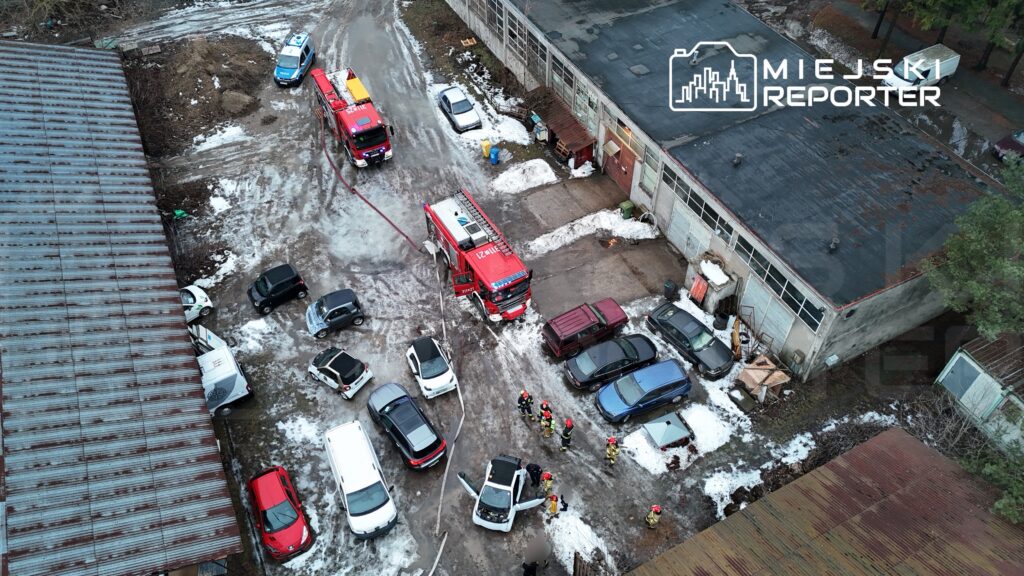 Grupa strażaków w kaskach pracuje przy pojeździe w zarośniętym terenie, obok wozów strażackich.