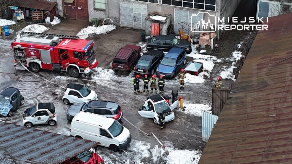 Grupa strażaków w hełmach rozmawia obok białego samochodu z otwartą maską na zaśnieżonym parkingu. W tle widać woz strażacki.