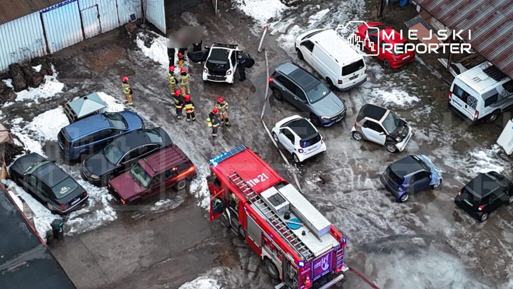 Grupa strażaków w kaskach pracuje przy uszkodzonym samochodzie na parkingu, obok stoi wóz strażacki.