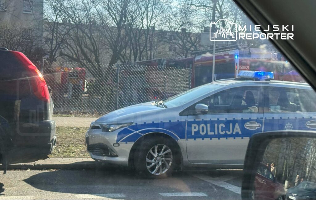 Policyjny radiowóz z niebieską sygnalizacją zaparkowany obok czarnego SUV-a, w tle widać wóz strażacki.