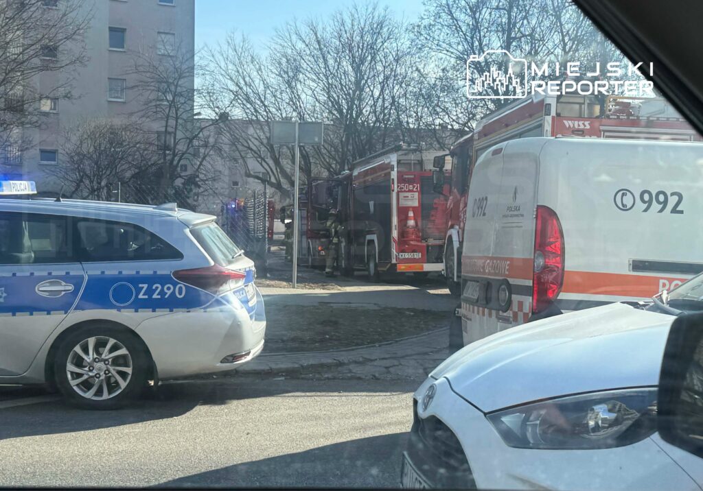 Na ulicy zaparkowane pojazdy służb ratunkowych, w tym radiowóz policji oraz samochody straży pożarnej i pogotowia gazowego.