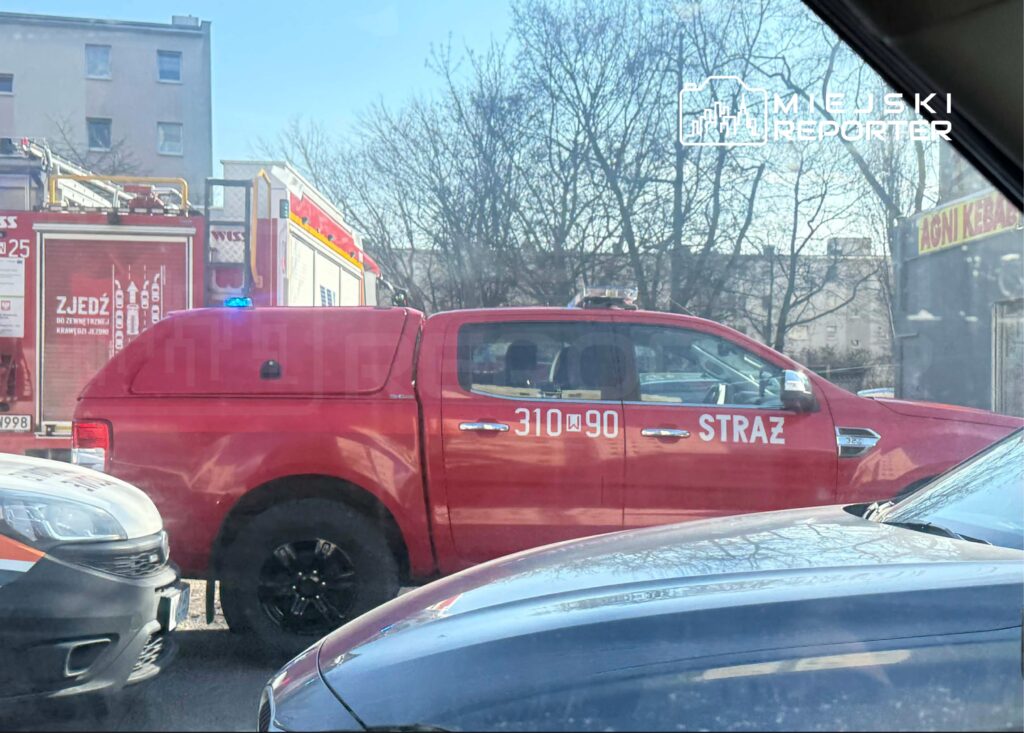 Czerwony samochód strażacki z napisem "STRAŻ" stoi na ulicy, obok widoczna ciężarówka strażacka.