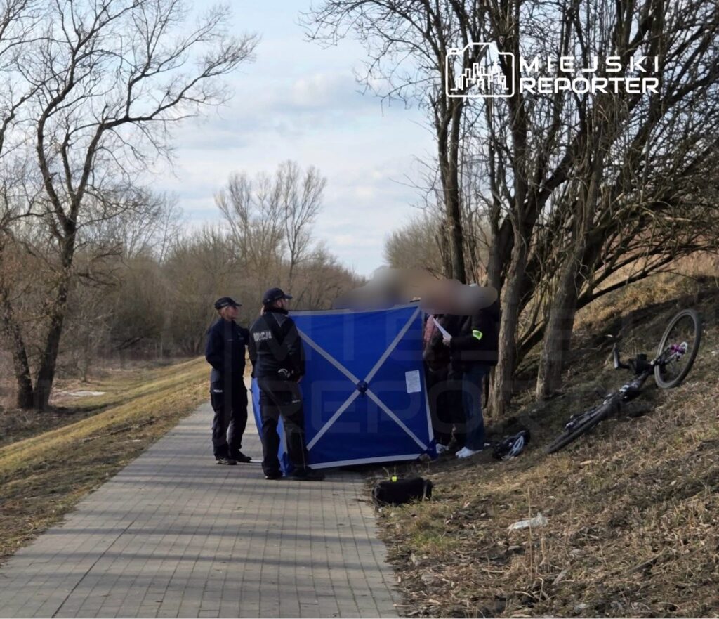 Policjanci rozmawiają obok niebieskiego namiotu wzdłuż ścieżki rowerowej, obok leżącego roweru.