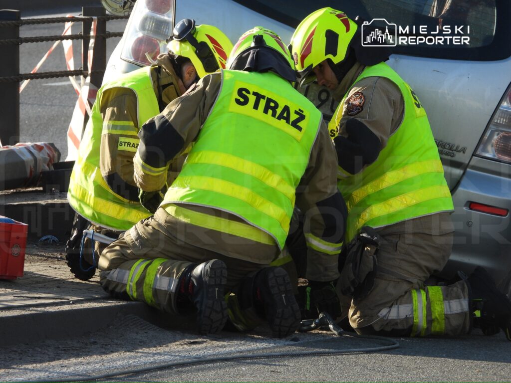 Czterech strażaków w odblaskowych kamizelkach pracuje nad usunięciem uszkodzonego pojazdu na drodze.