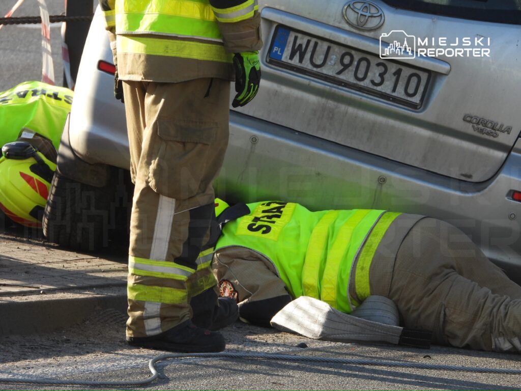 Strażacy w kamizelkach odblaskowych sprawdzają osobę leżącą na ziemi obok samochodu, który jest częściowo uniesiony.