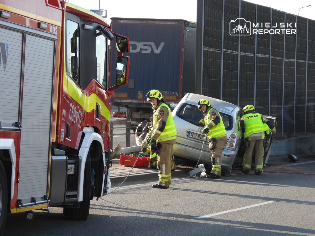Strażacy w żółtych kaskach i kamizelkach pracują przy przewróconym samochodzie osobowym obok drogi.