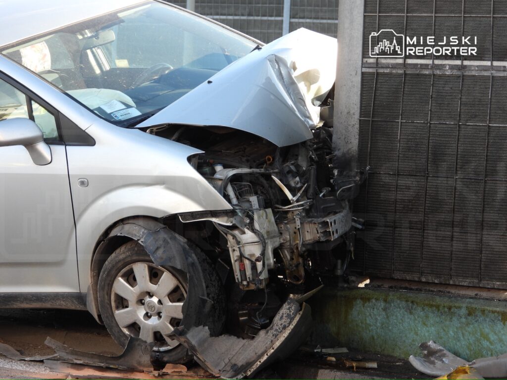 Uszkodzony srebrny samochód osobowy z wgniecionym przodem uderzył w stalowy słup, leżący na poboczu.