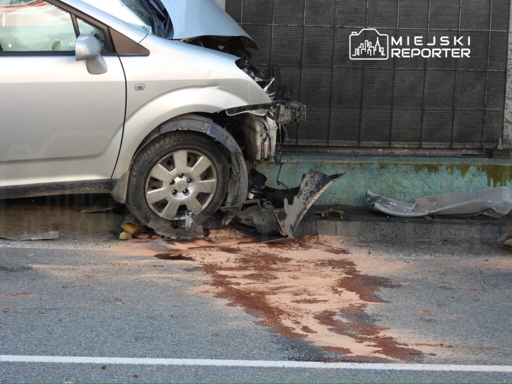 Uszkodzony samochód osobowy z oderwaną przednią częścią i wyciekającym płynem na nawierzchni ulicy.