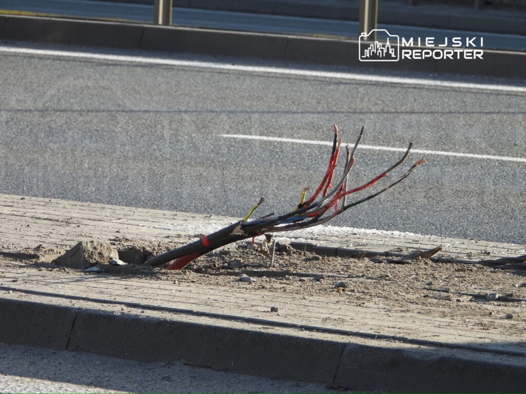 Uszkodzone kable wystają z rozkopanej nawierzchni chodnika przy ulicy, obok jezdni.